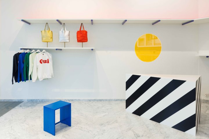 Detailansicht der Verkaufstheke im «La Garçonne» Store: Eine weisse Theke mit markanten schwarzen Diagonalstreifen auf Marmorboden. Darüber hängt eine kreisrunde, gelb getönte Glasscheibe als Trennelement. Im Hintergrund sind bunte Taschen und Kleidung an minimalistischen weissen Regalen sichtbar.