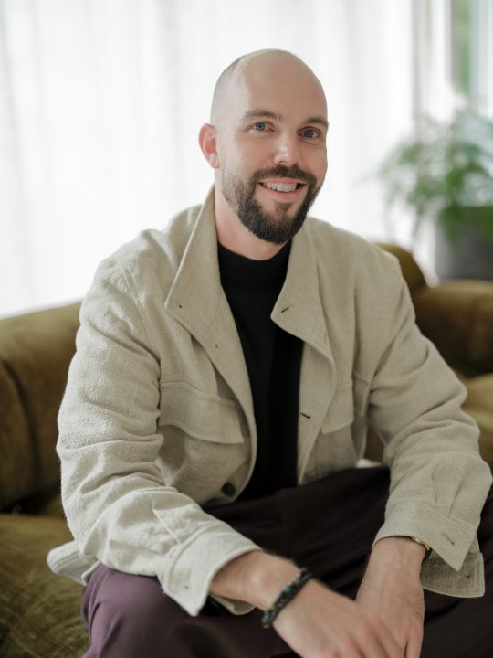 Porträt von Fabian Messer: Ein lächelnder Mann mit Glatze und dunklem Vollbart, der in einer hellen, modernen Umgebung auf einem olivgrünen Sofa sitzt.