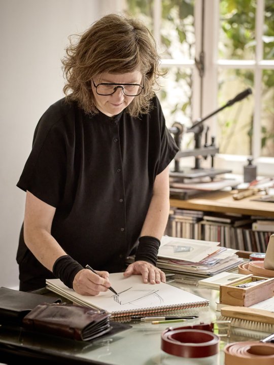 Designerin zeichnet eine Skizze in ein Skizzenbuch an einem Arbeitstisch, umgeben von Leder, Werkzeugen und Arbeitsmaterialien.