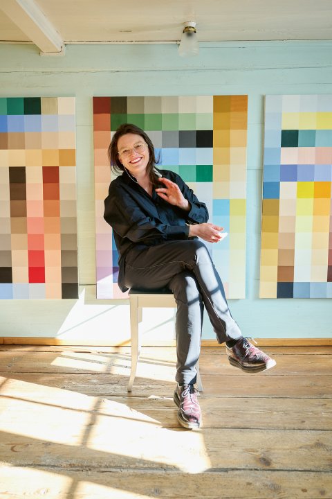 Eine Frau sitzt mit übereinandergeschlagenen Beinen lachend auf einem Stuhl. Im Hintergrund Bilder aus Farbquadraten.